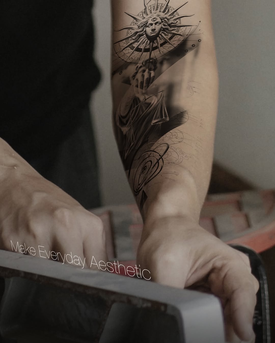 Black and grey tattoo of a sun-faced figure with radiating spikes and draped fabric on the inner forearm, featuring detailed shading and decorative line work below.
