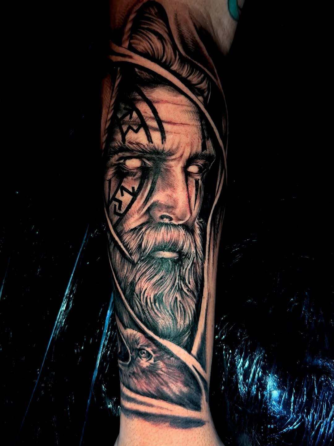 Bearded man's portrait with tribal face markings and geometric patterns, rendered in monochromatic black and grey on forearm.