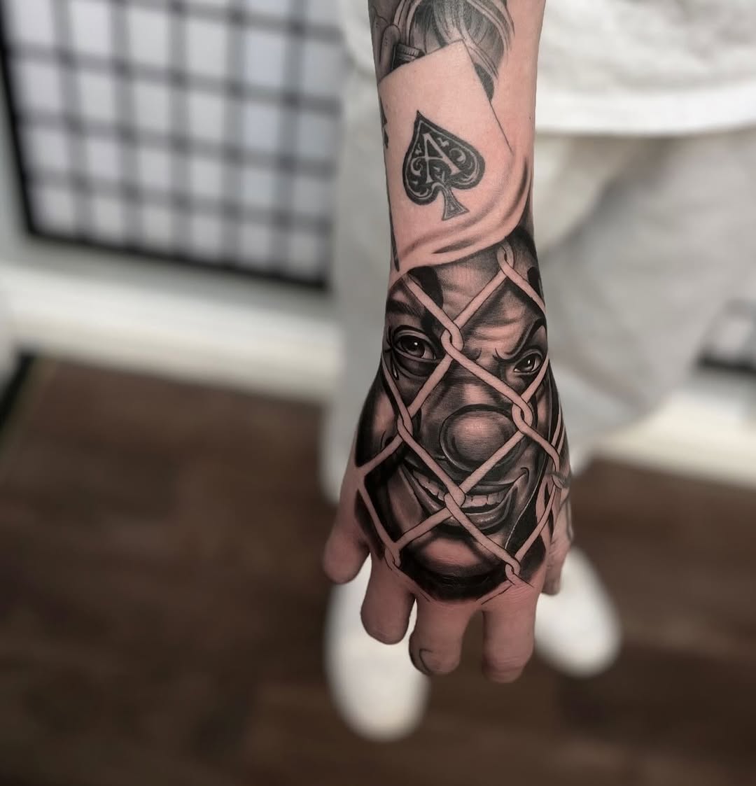 Black and grey portrait tattoo of a face with chains wrapped across it on the hand, with a spade symbol visible on the forearm above.