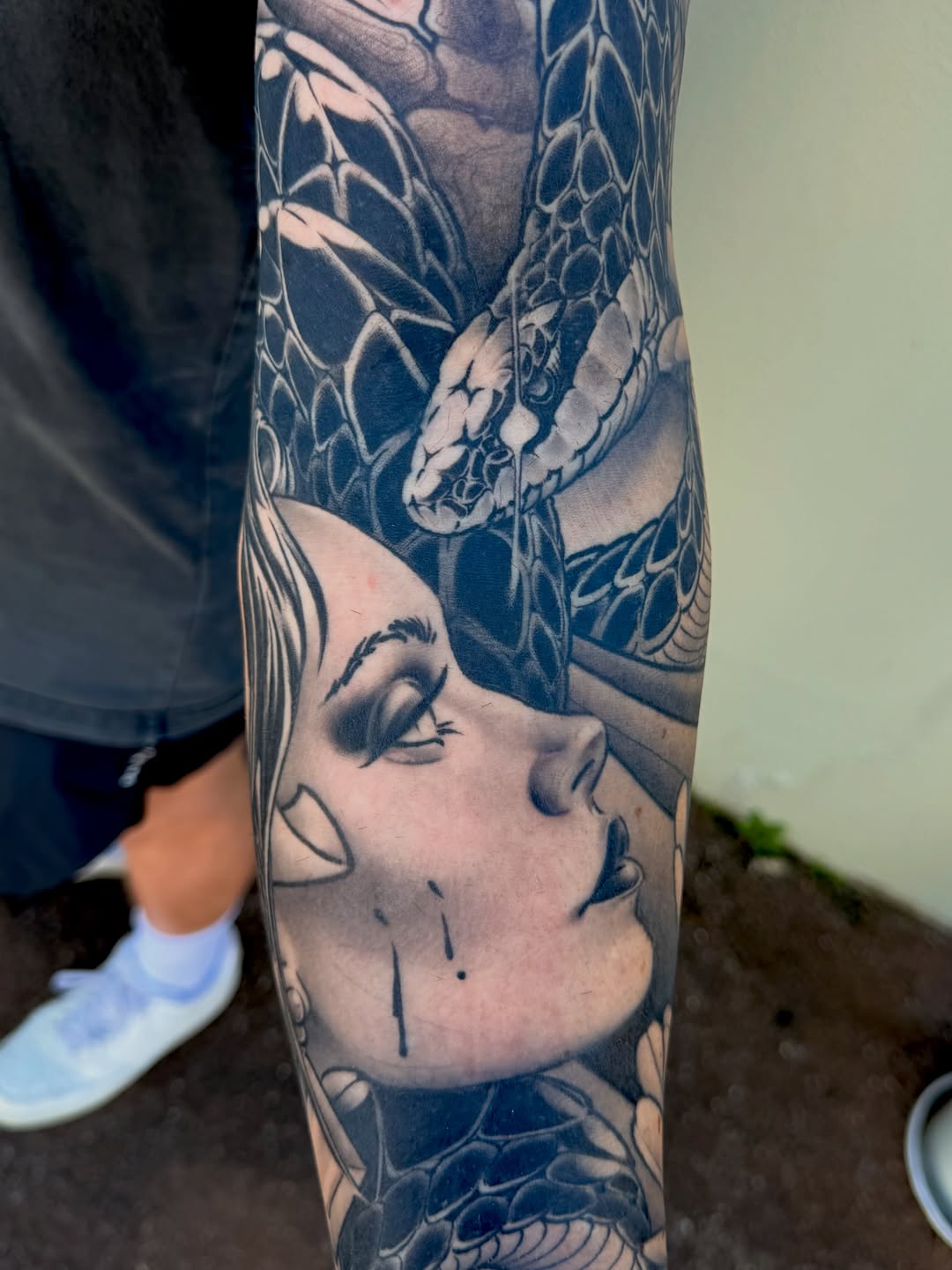 Black and grey sleeve tattoo featuring a woman's face with closed eyes and detailed dragon scales wrapping around the upper arm.