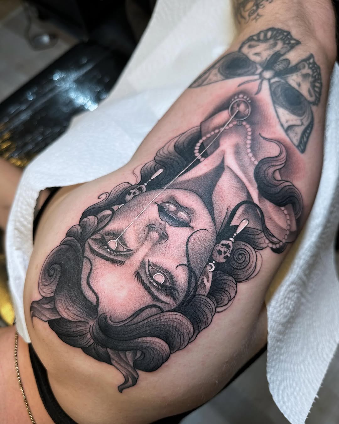 Black and grey upper arm tattoo of a woman's face with flowing wavy hair, a pearl-headed pin, and a moth above.