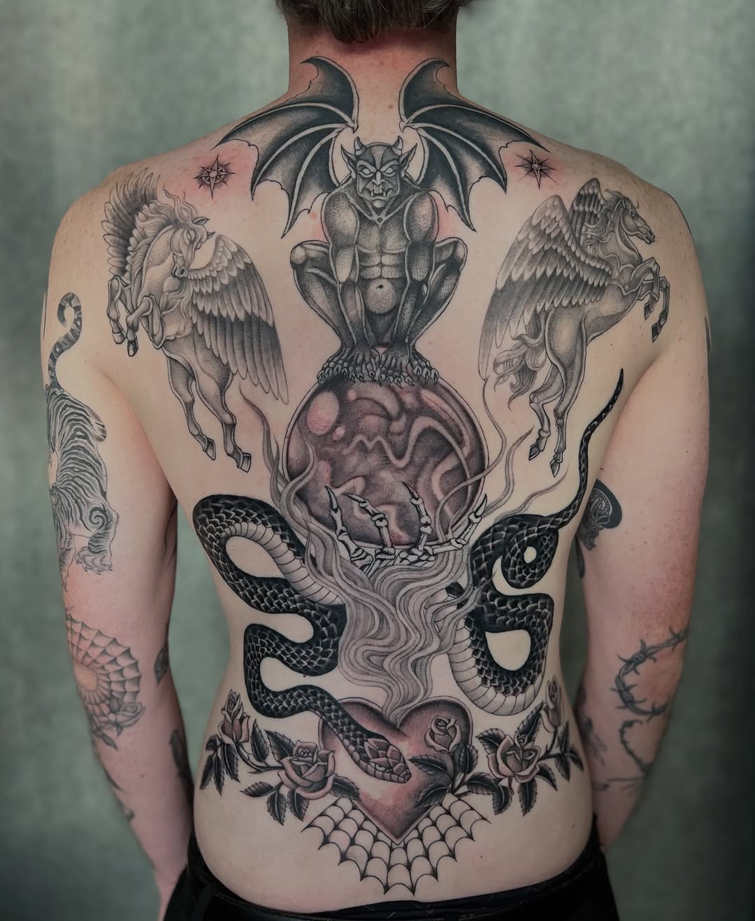 Full back tattoo featuring a gargoyle atop a globe, winged horses, snakes, roses, and a heart, in black and grey shading.