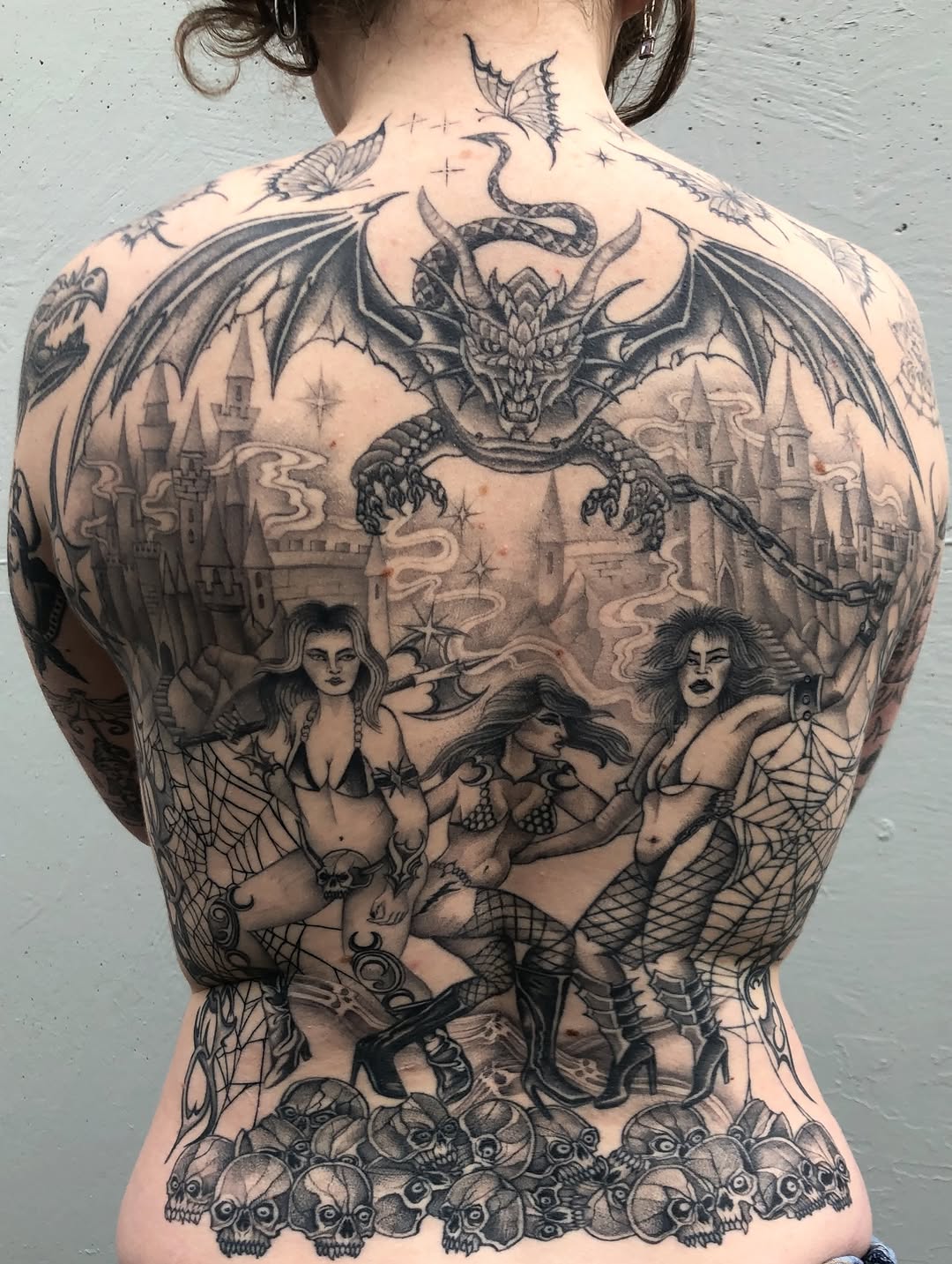 Full back tattoo featuring a dragon, gothic castle, three pin-up women, skulls, and butterflies in black and grey shading.