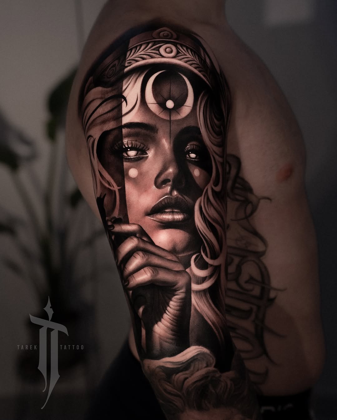 Black and grey portrait tattoo of a woman's face with ornamental headpiece and third eye symbol on upper arm.
