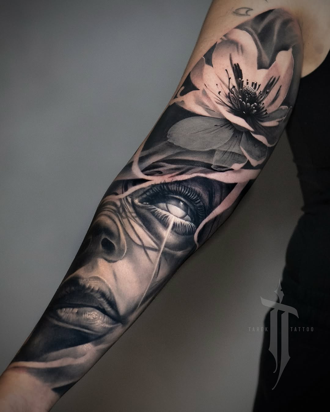 Black and grey realistic portrait tattoo on forearm featuring a man's face with a flower blooming from his eye.