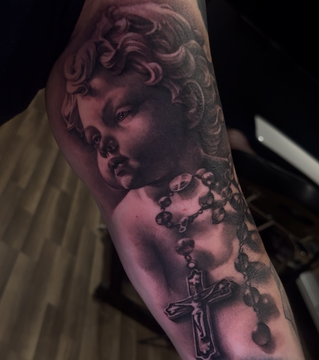 Black and grey portrait tattoo of a classical cherub's face with curled hair and a cross with rosary beads on the upper arm.