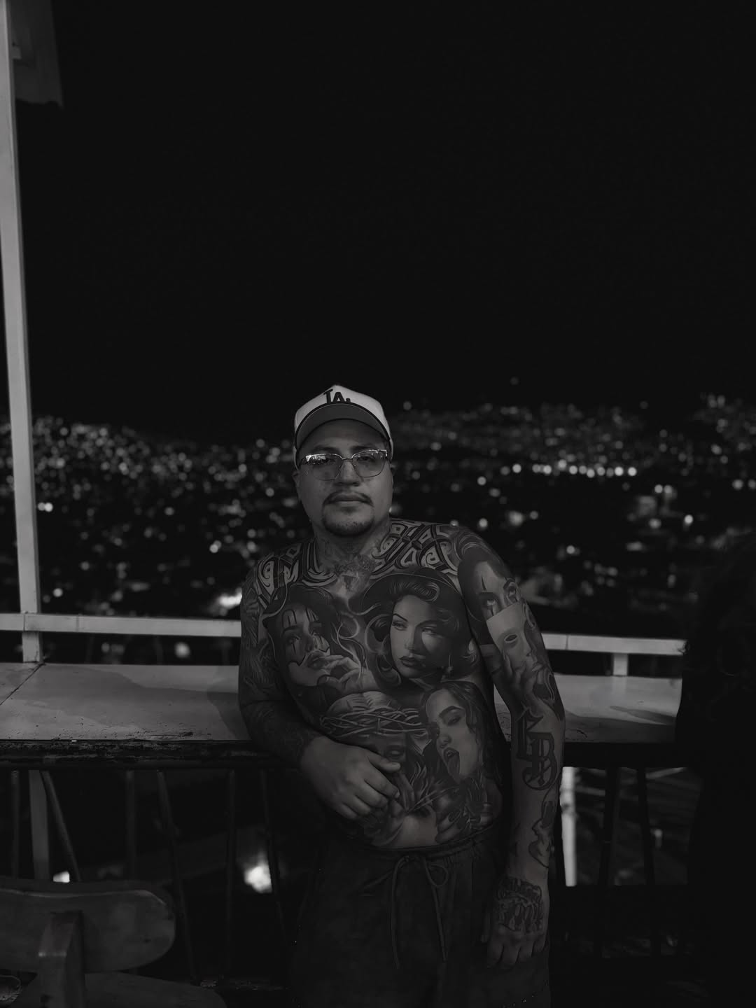Black and grey portrait tattoos covering the chest and arms of a man wearing a cap and glasses against a city skyline.