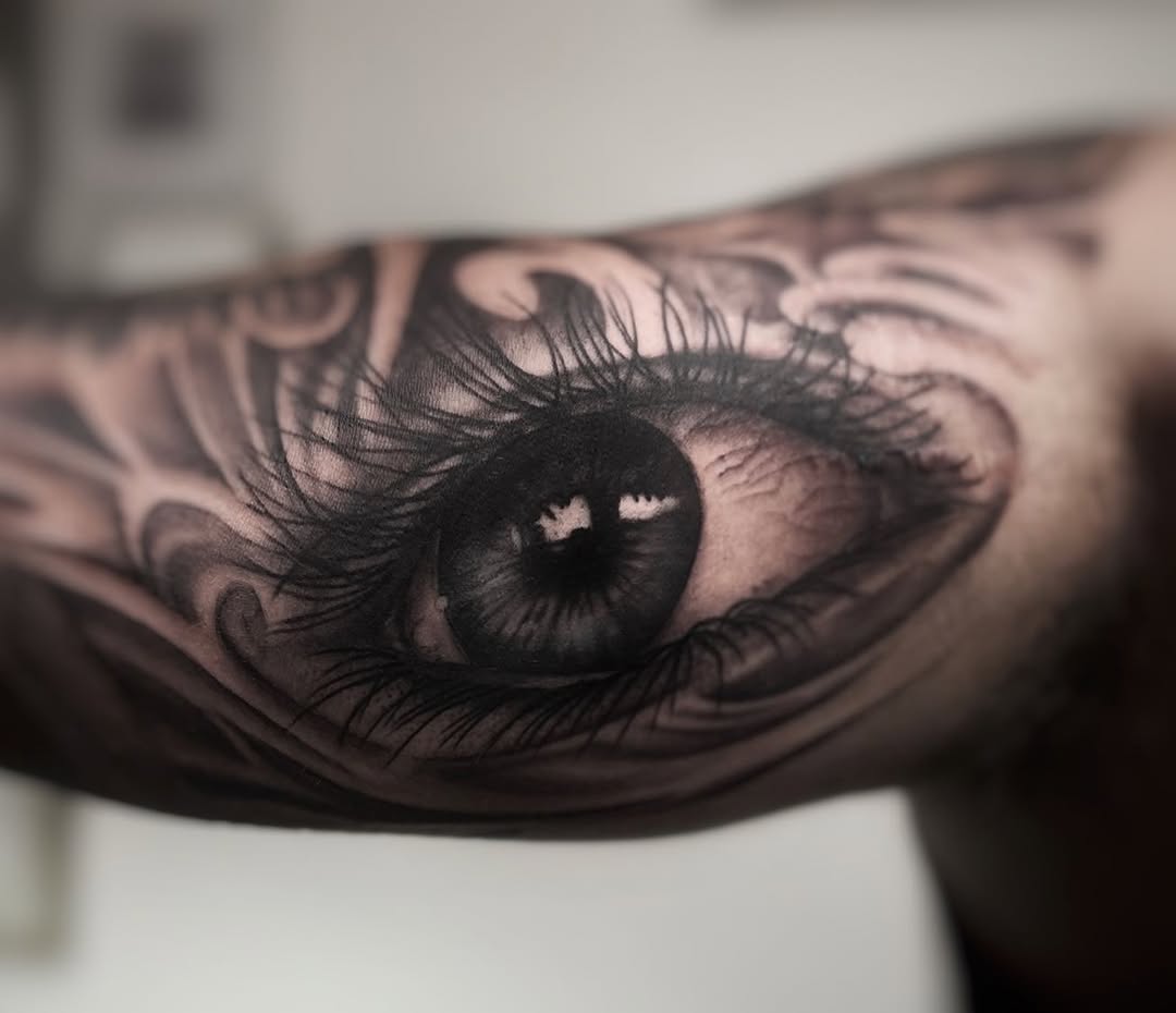 Realistic black and grey eye tattoo with detailed lashes and iris on forearm, featuring ornamental line work surrounding it.