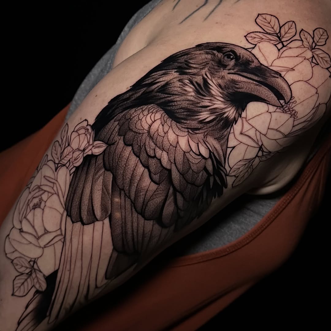 Raven with detailed feathers among linework peonies and leaves, covering an upper arm, in shades of black and grey.