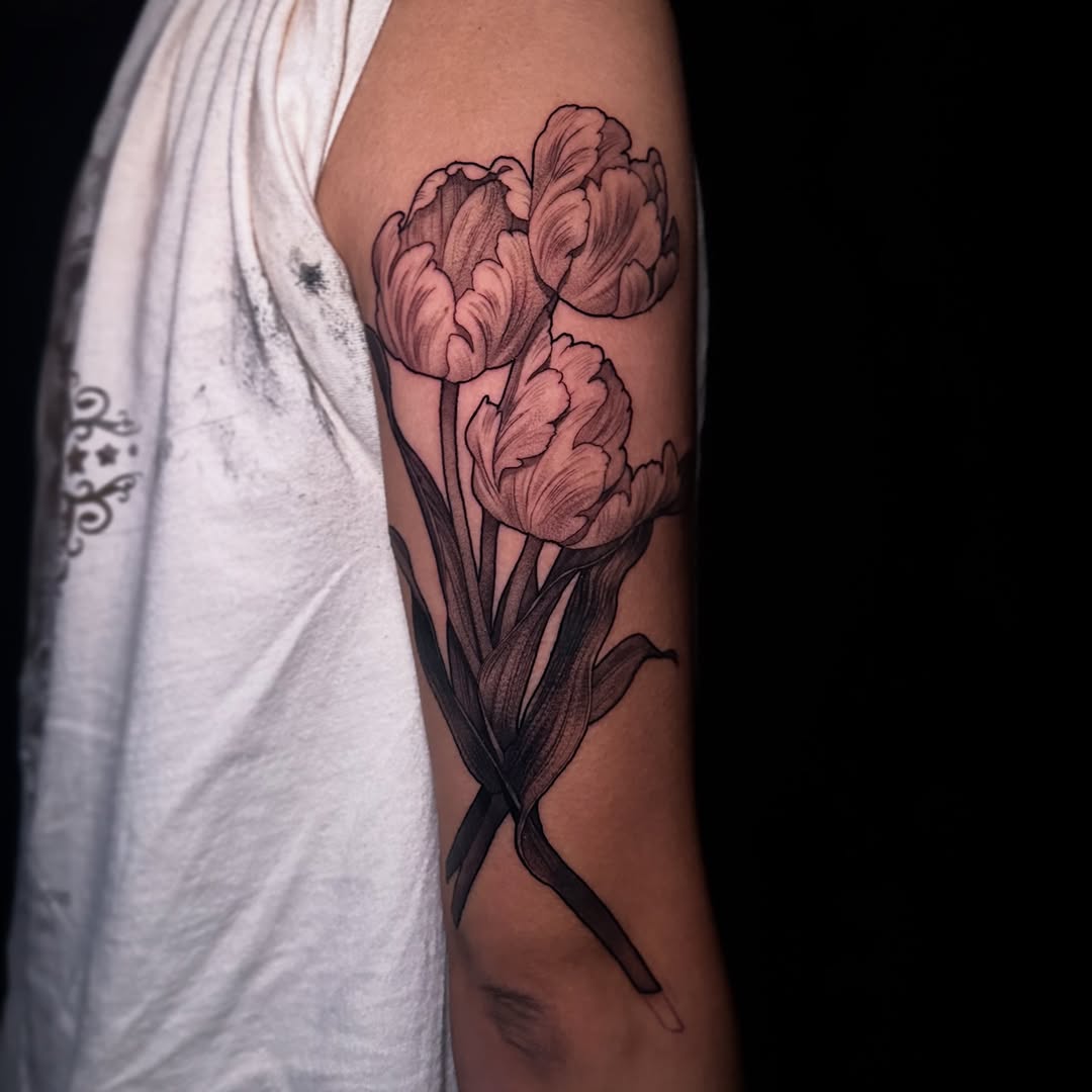 Three parrot tulips with flowing leaves on an upper arm, softly shaded in black and grey.