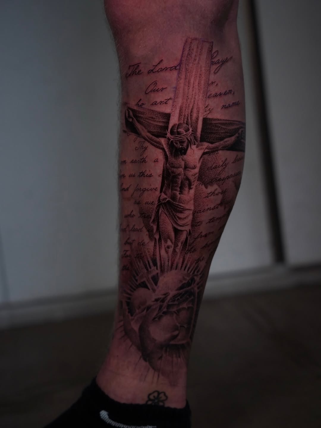 Black and grey crucifix tattoo with religious script text on calf, featuring detailed shading and a glowing halo effect.