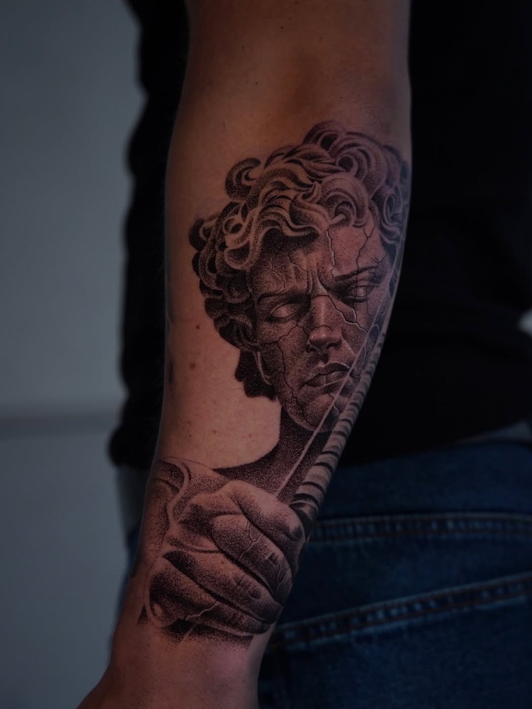 Black and grey classical male bust portrait tattoo on forearm with curly hair and draped clothing detail.
