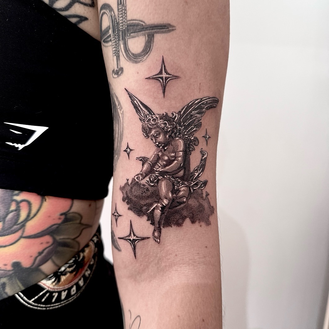 Black and grey tattoo of a winged cherub sitting on a cloud surrounded by four-point stars, placed on the inner forearm.