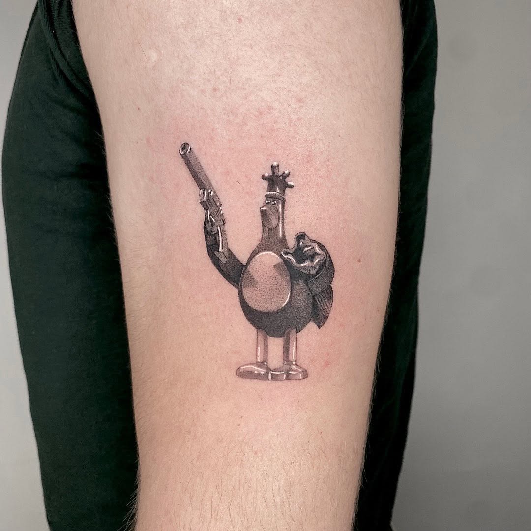 Black and grey tattoo of a cartoon bird wearing a crown holding a revolver, on upper arm.