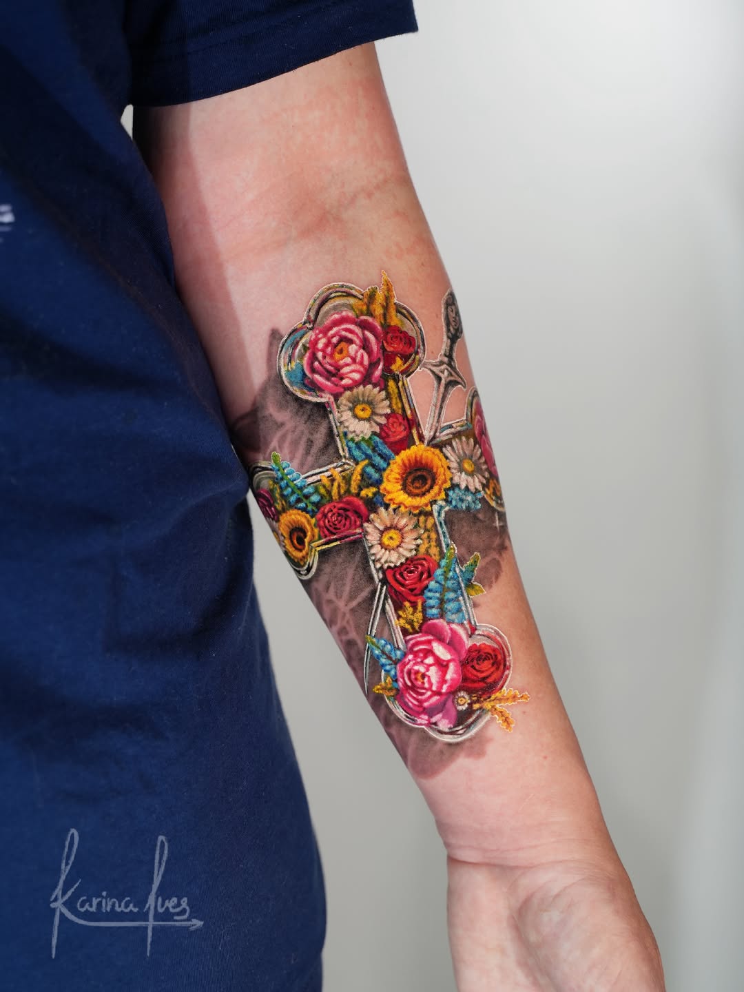 Floral cross tattoo with roses, sunflowers, and peonies on forearm with ornamental frame details.