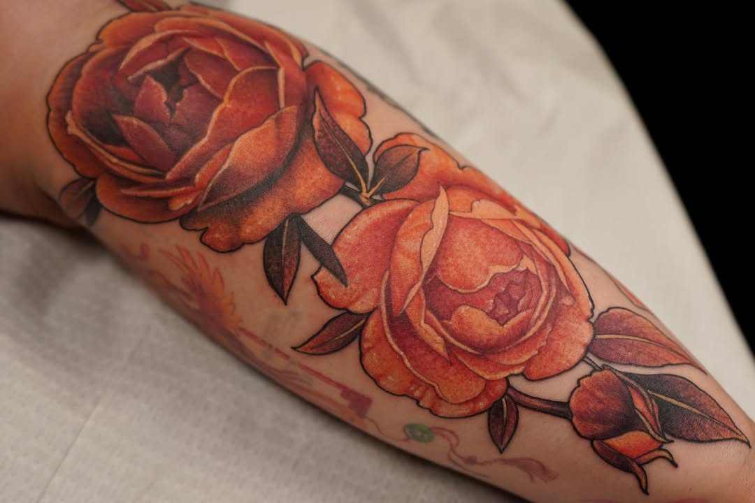 Three red and orange peonies with dark green leaves on the forearm, displaying detailed shading and layered petals.