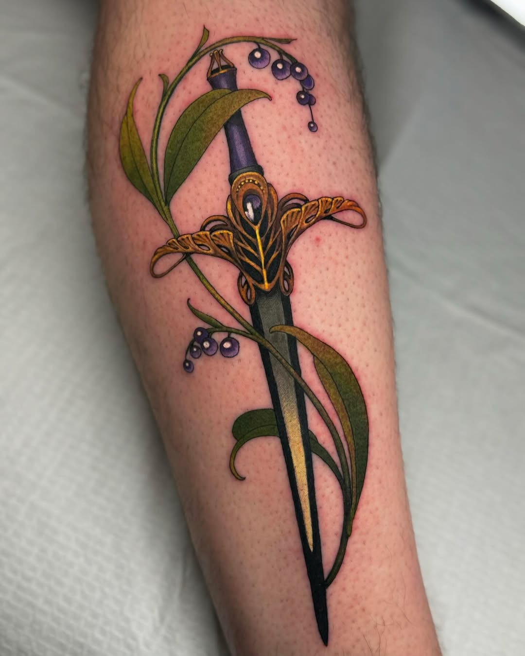 A decorative sword with ornate gold crossguard wrapped in green leafy vines bearing purple berries on the forearm.