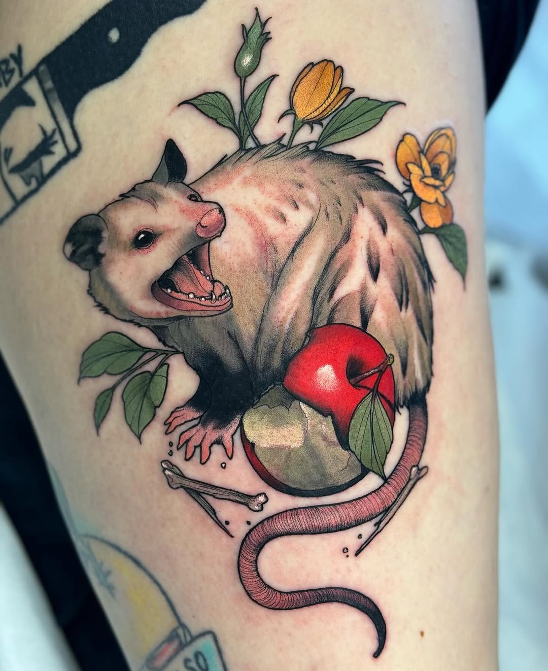 A possum with open mouth surrounded by colorful flowers, a red apple, bones, and a coiled snake on the upper arm.