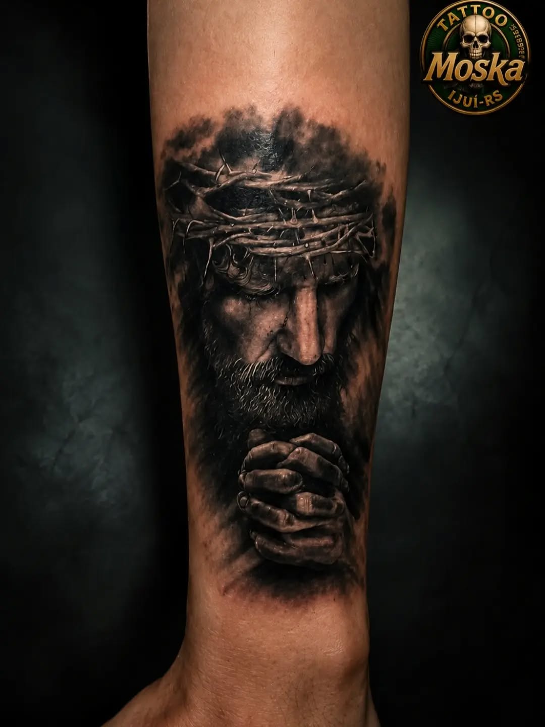 Portrait of bearded man wearing crown of thorns with clasped hands, rendered in monochrome black and grey on forearm.