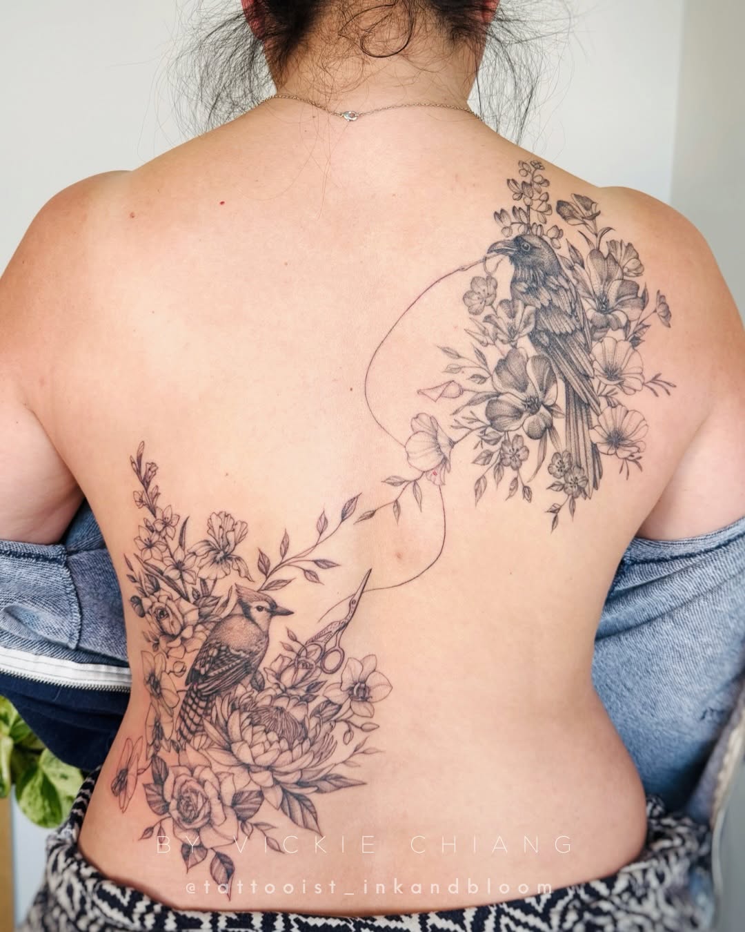 Back tattoo featuring two birds surrounded by flowering botanicals connected by flowing curved linework across the spine.