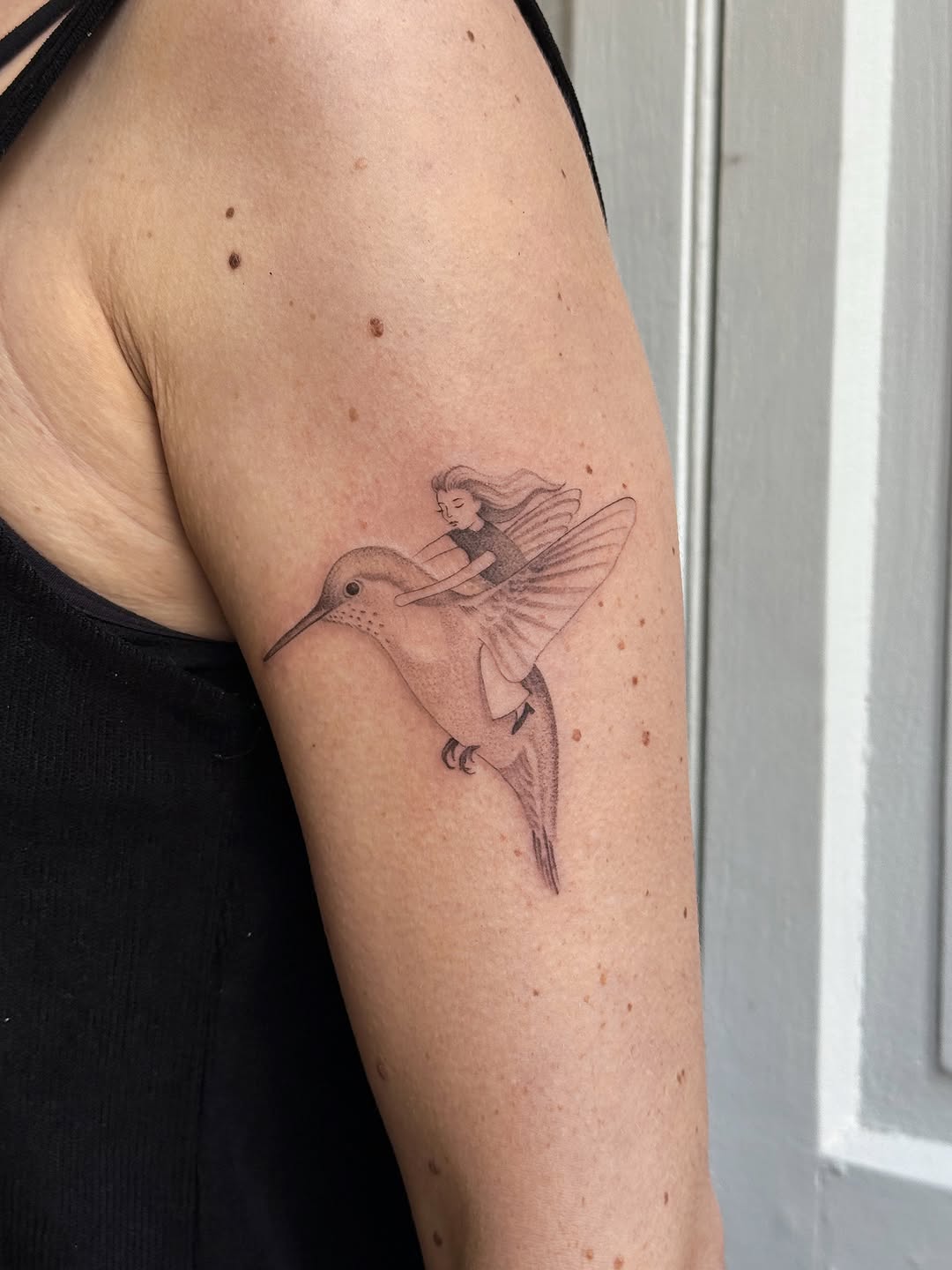 Delicate hummingbird with a small fairy figure seated on its back, rendered in thin linework on the upper arm.