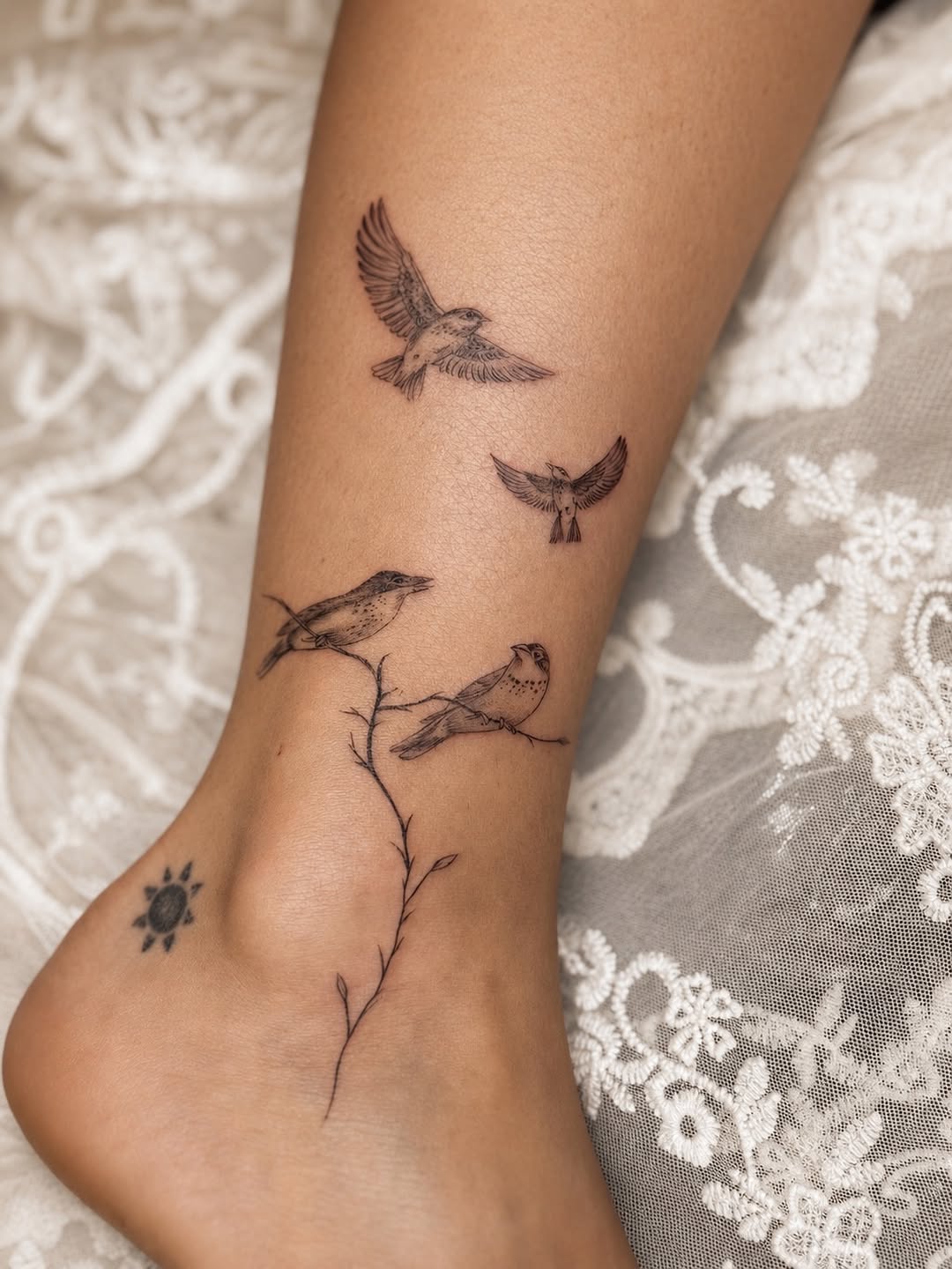 Delicate fine-line birds in flight above a thin branch with small leaves on the inner forearm and ankle.