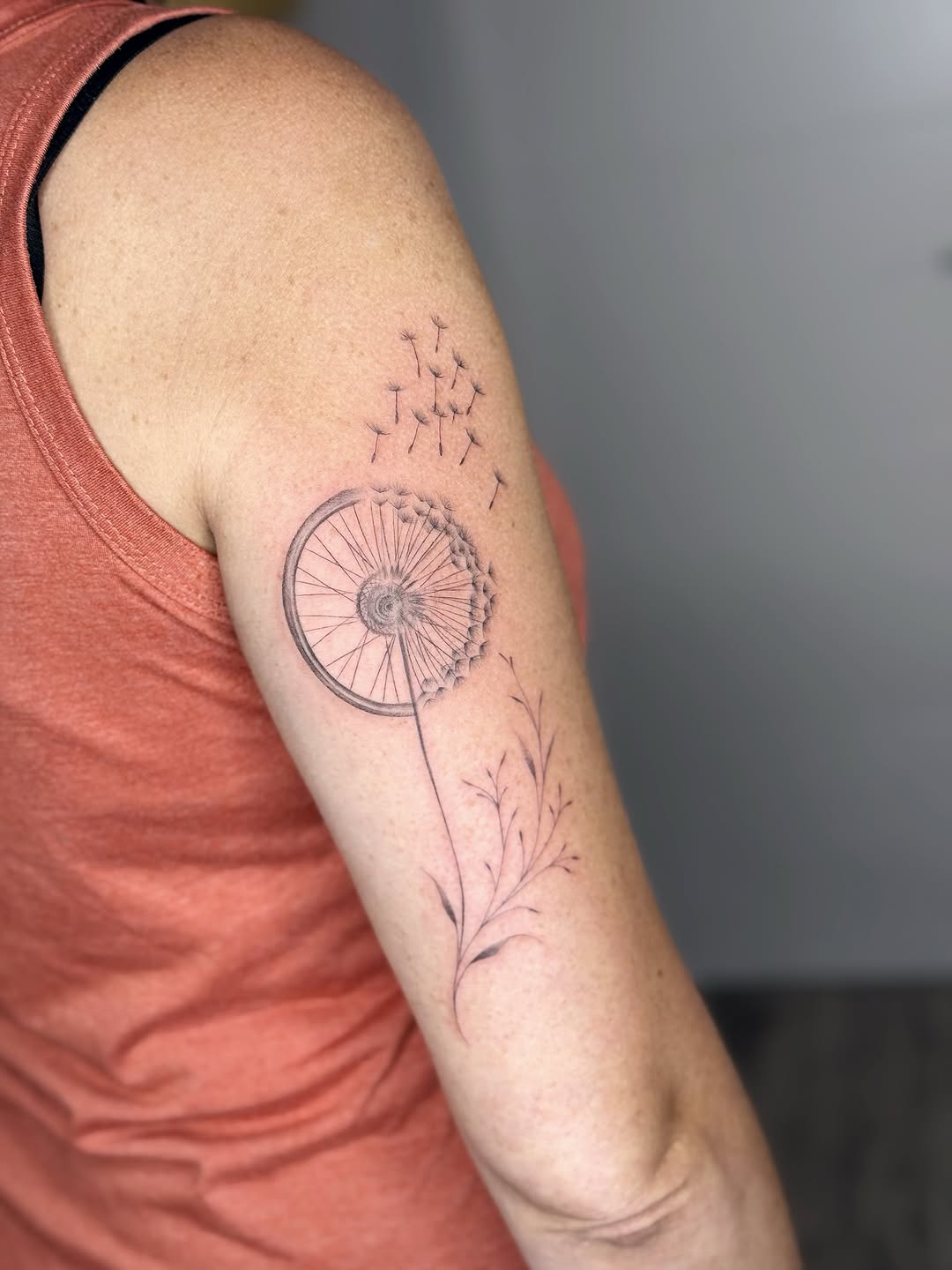 Delicate line work tattoo featuring a bicycle wheel with flowering stem and scattered seeds on the upper arm.