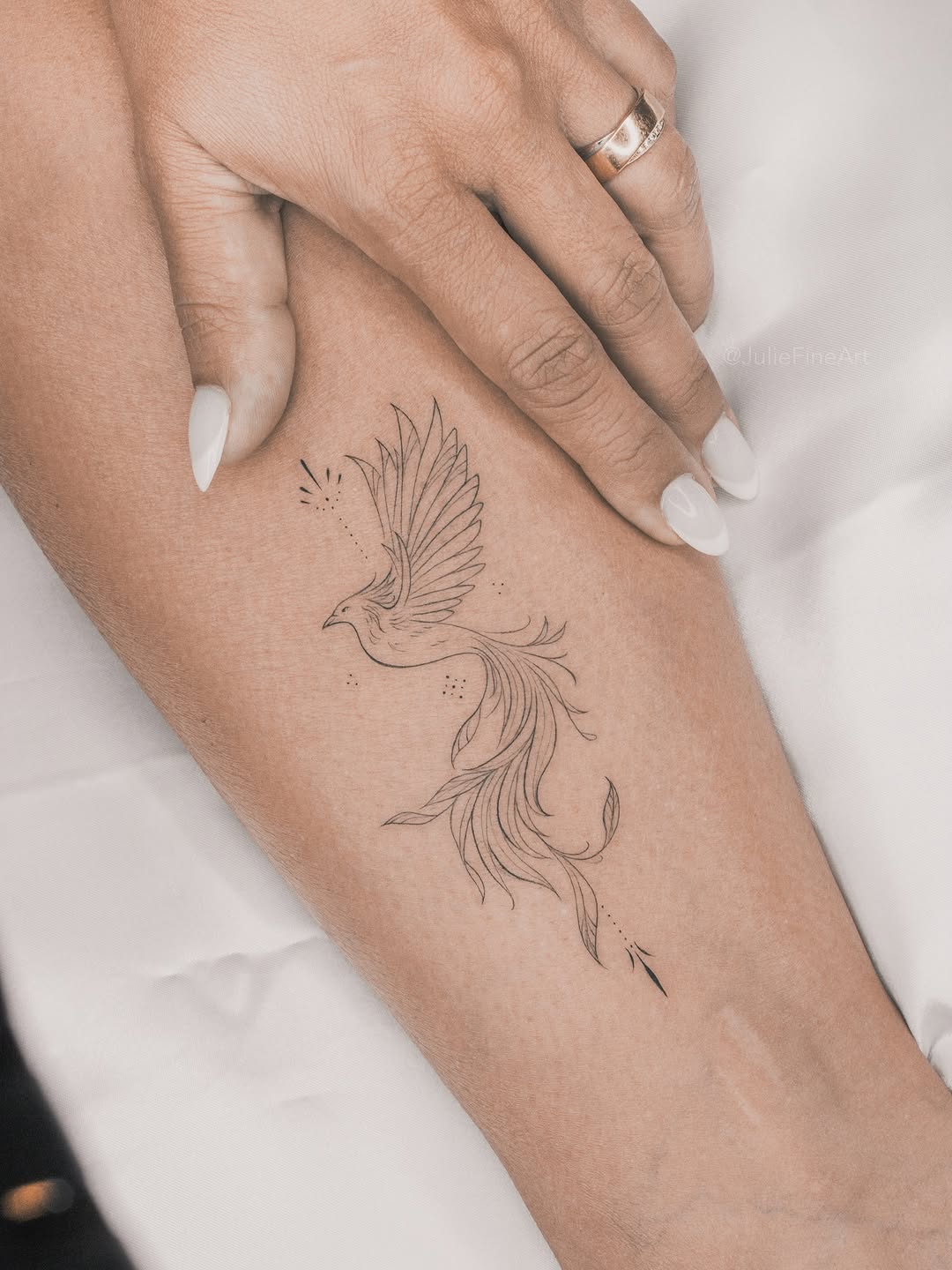 Delicate phoenix bird with flowing feathers and scattered dot accents on the inner forearm, rendered in thin black linework.