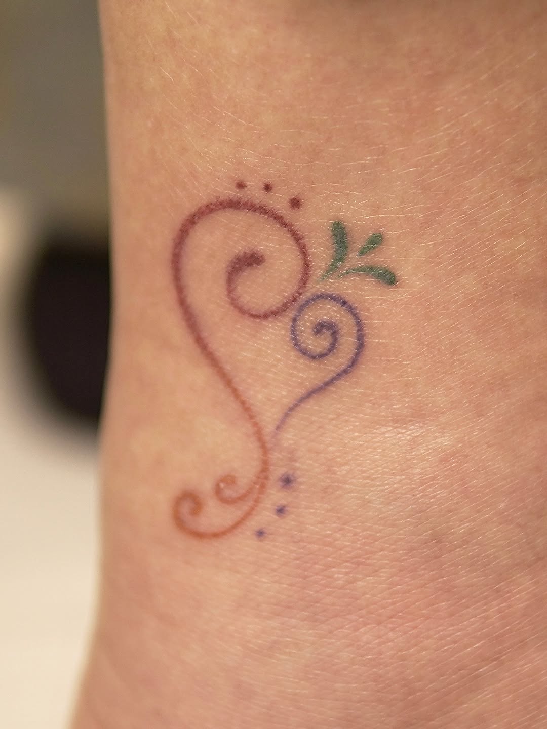 Delicate vine with two spiral flowers and small dots, rendered in thin purple and green linework on the inner forearm.
