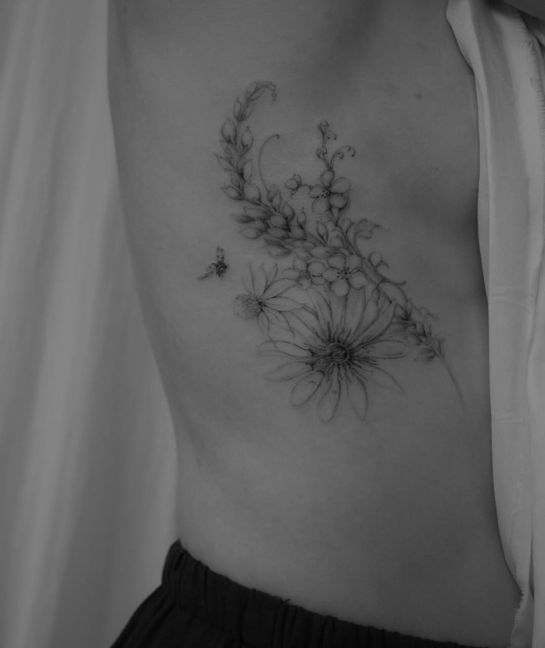 Delicate fine-line floral composition featuring a large daisy with accompanying botanical branches and smaller flowers on the ribcage.