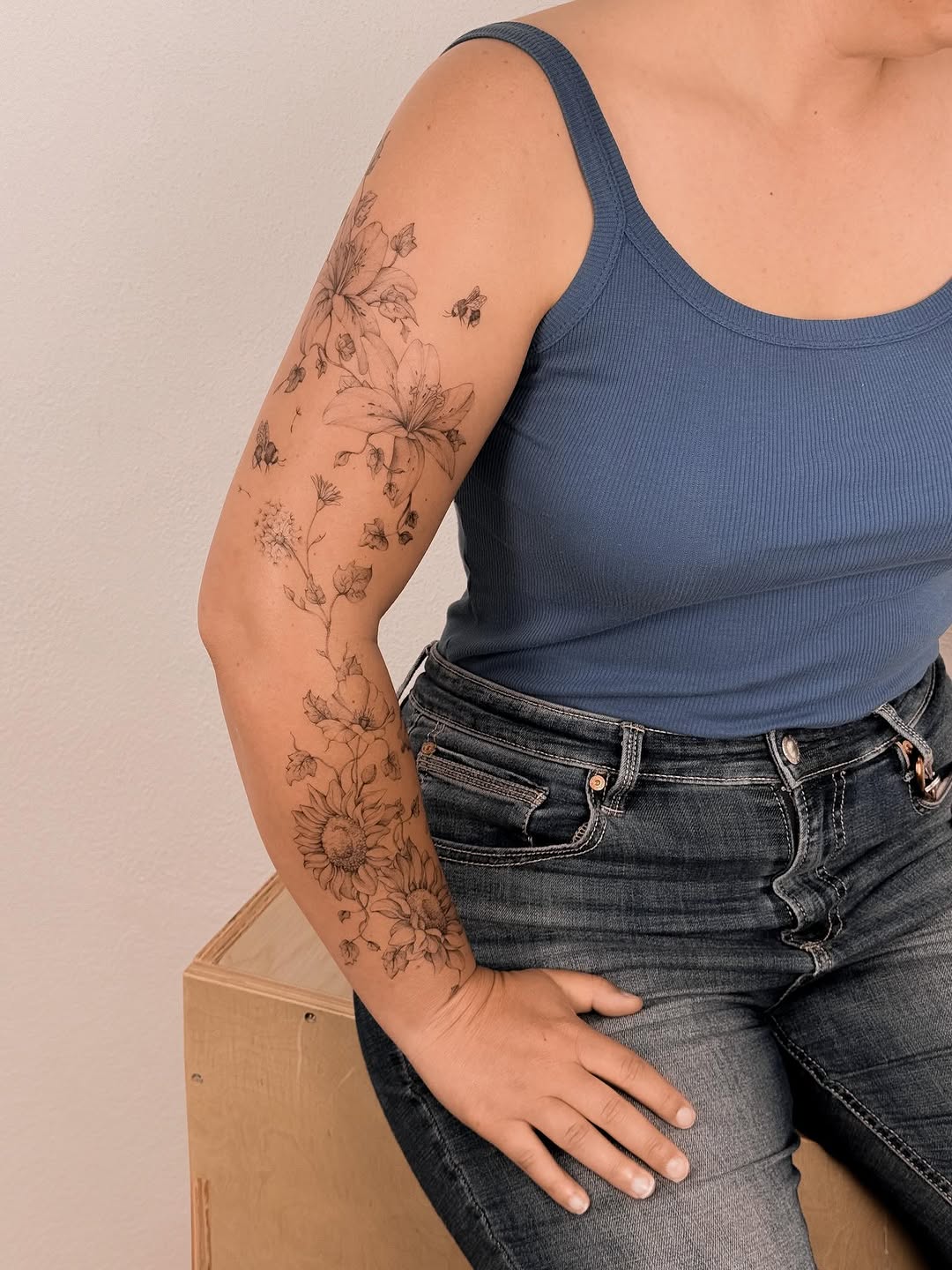 Delicate botanical sleeve tattoo featuring sunflowers, daisies, and small bees in thin linework extending down the upper arm.