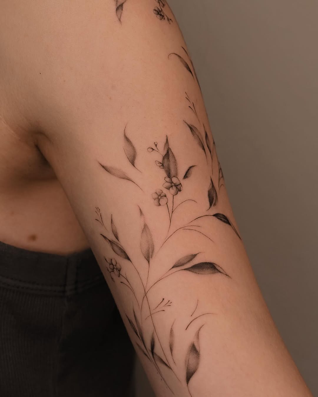 Delicate botanical tattoo with thin-lined flowering stems and leaves wrapping around the upper arm.