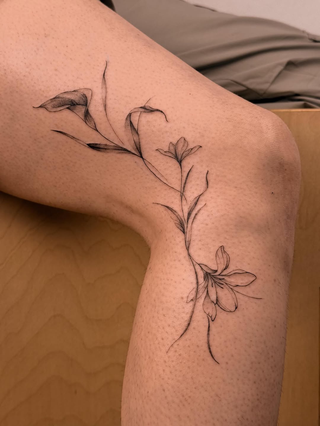 Delicate botanical line tattoo featuring flowering stems with buds and blossoms on the thigh.