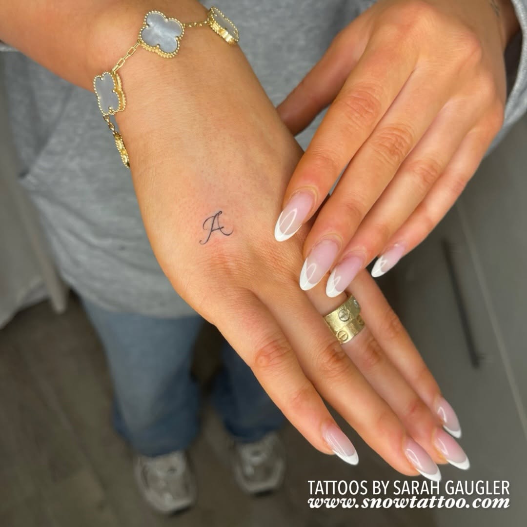 Delicate cursive letter 'A' tattoo on the hand between thumb and index finger.