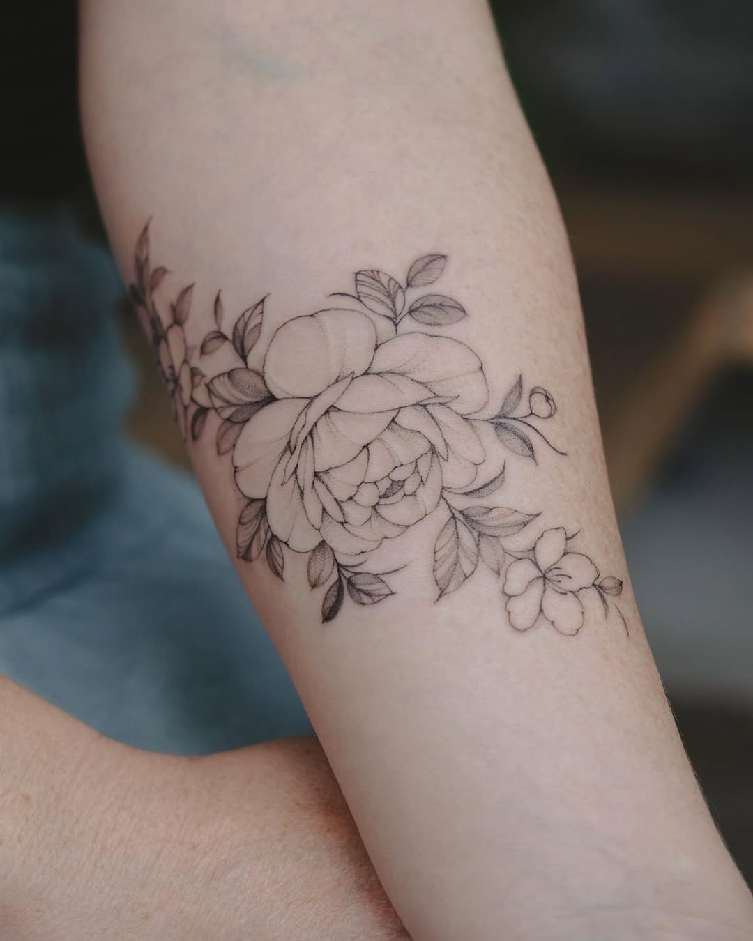 Delicate line-drawn floral composition featuring a large peony surrounded by smaller blooms and foliage on the forearm.