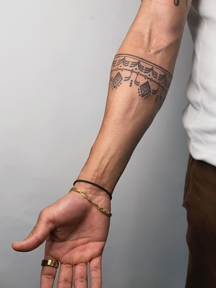Ornamental armband tattoo with scalloped lace pattern and hanging geometric pendants on forearm.