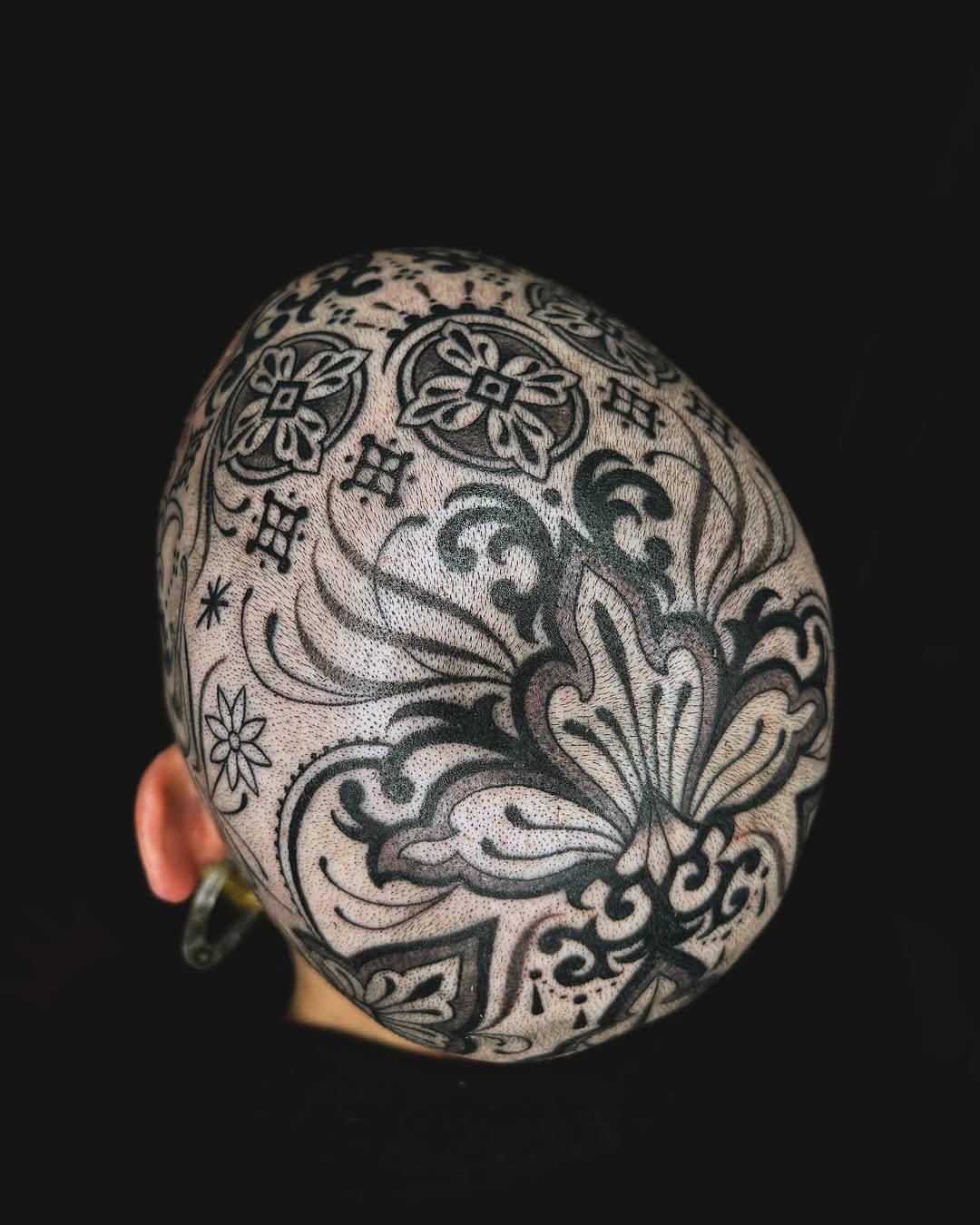 Intricate ornamental floral and scrollwork pattern covering the back of a shaved head in black ink.