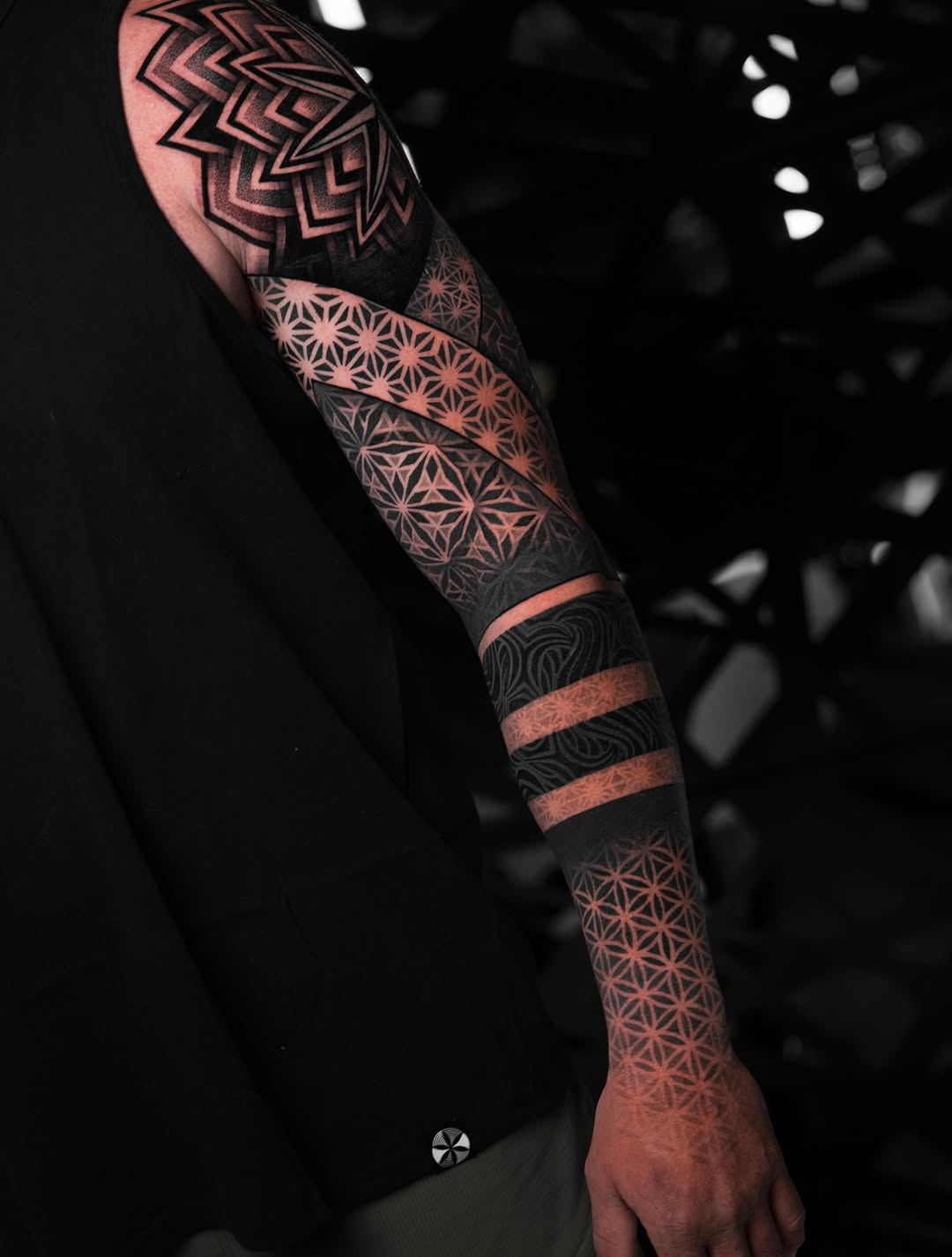Full-sleeve geometric tattoo featuring nested chevron patterns, tessellating star designs, and striped bands on arm.