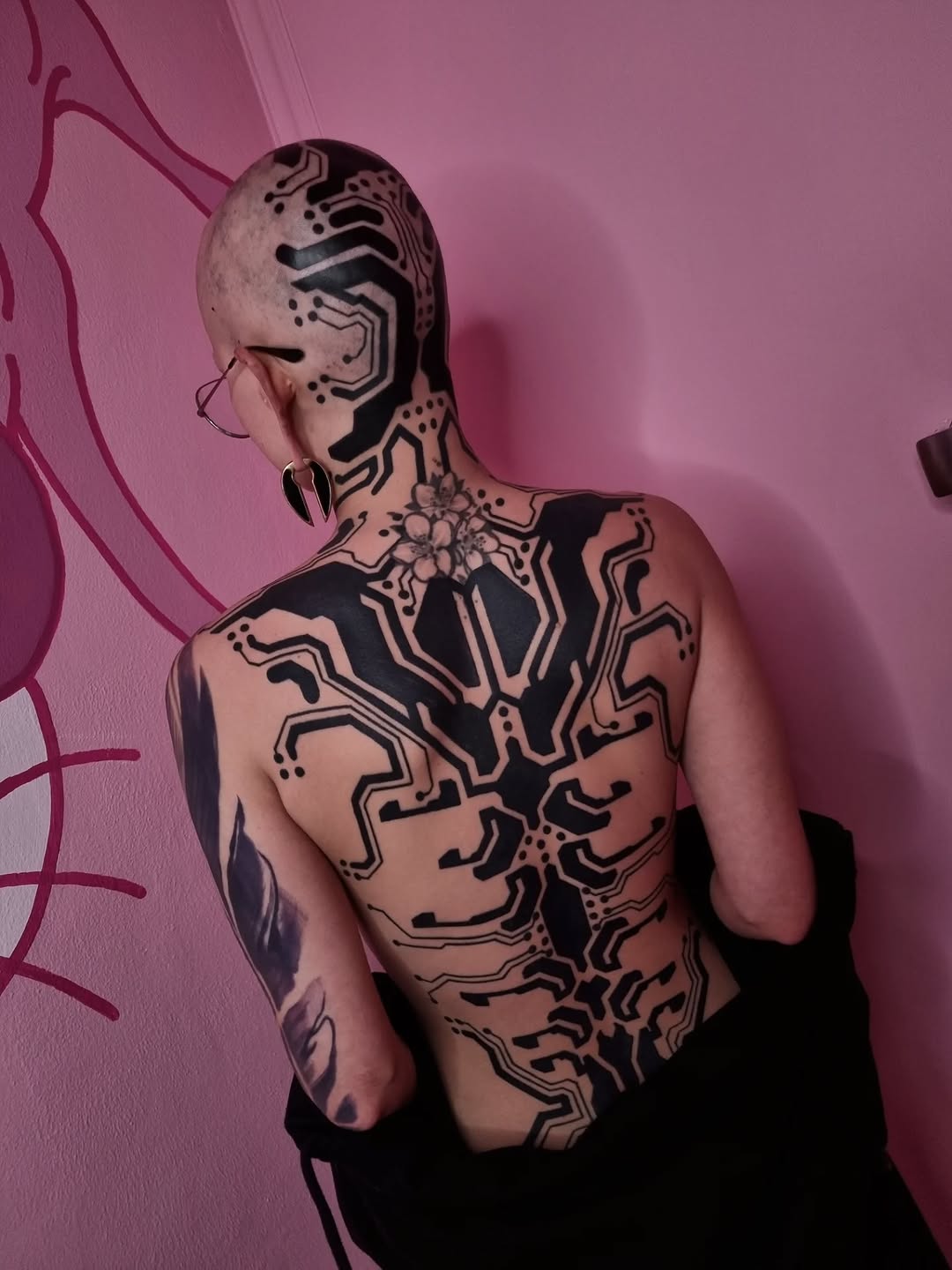 Full-body geometric tattoo with symmetrical line patterns, circuit-like designs, and ornamental detail covering torso and head.