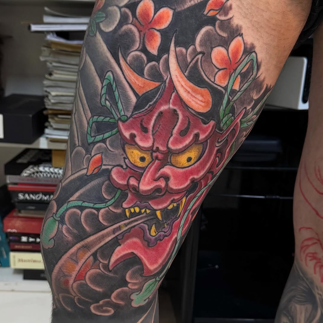 Red and black oni demon mask with orange flowers and green leaves on upper leg, traditional Japanese style.