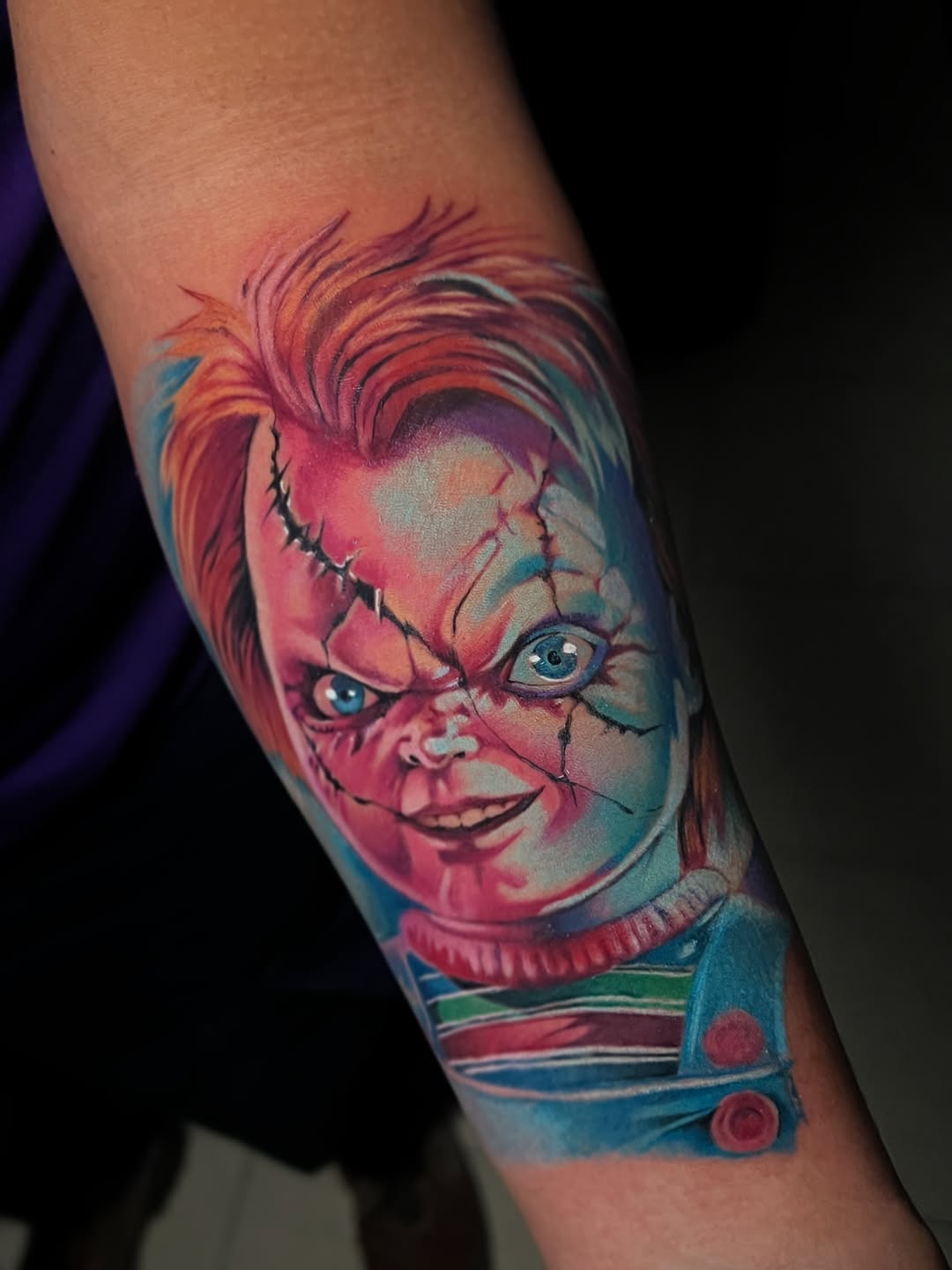 Chucky doll face with stitched seams, red and blue color palette, on forearm with clothing details visible.