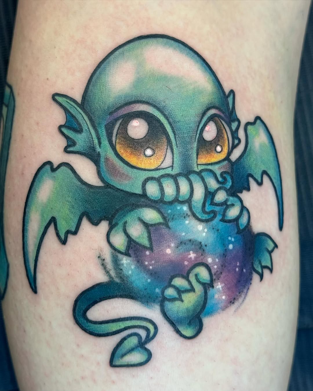 Cute teal creature with large golden eyes, bat wings, and sparkling belly on upper arm.