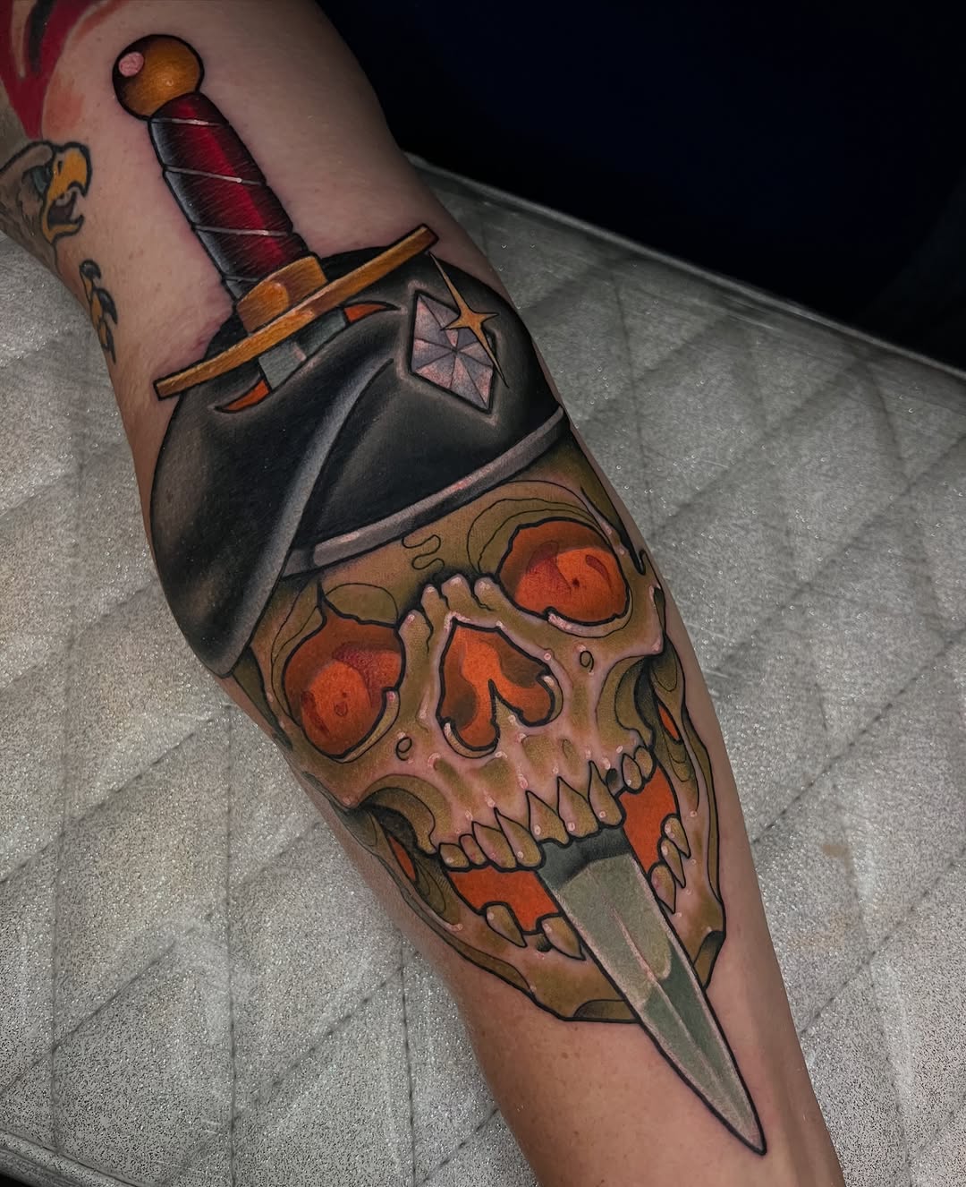 Skull with sword piercing through it, wearing black military helmet with red gem, on forearm with decorative shading.