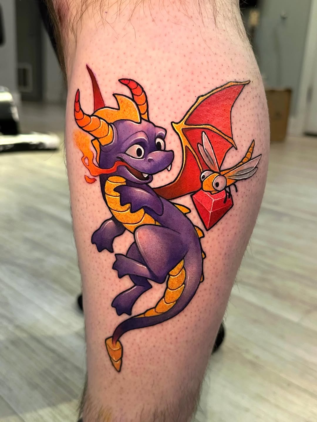 Purple dragon with orange horns and spikes, holding a red geometric crystal, on calf muscle.