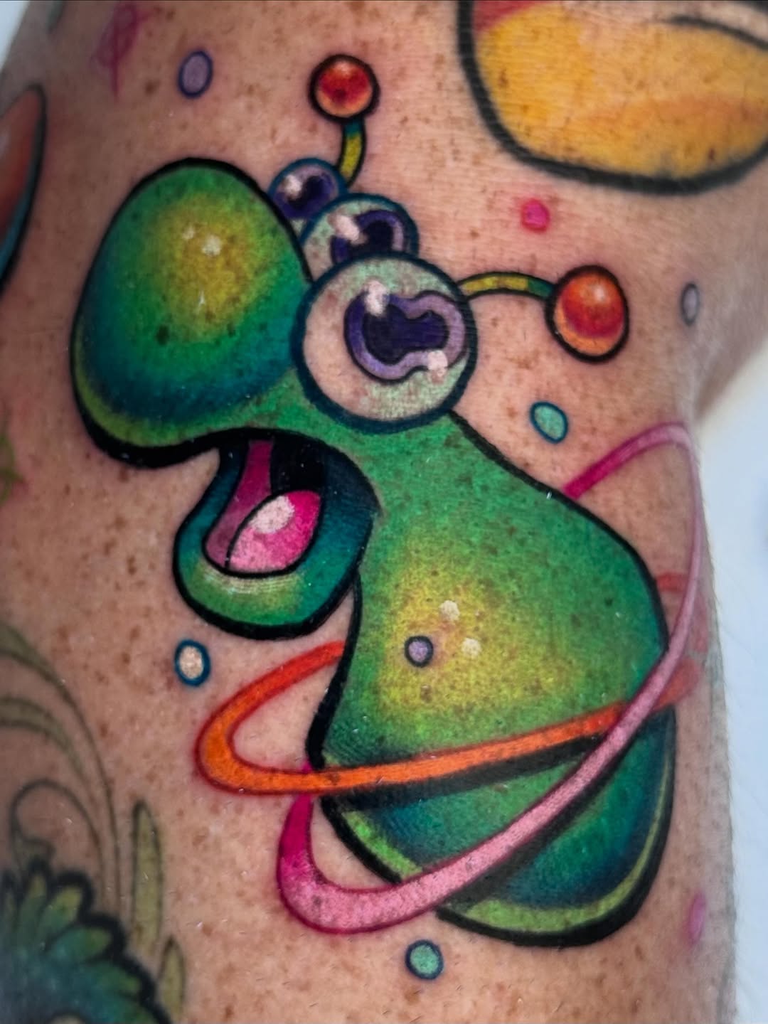 Green cartoon alien character with large expressive eyes, tentacles, and colorful planetary rings on upper arm.